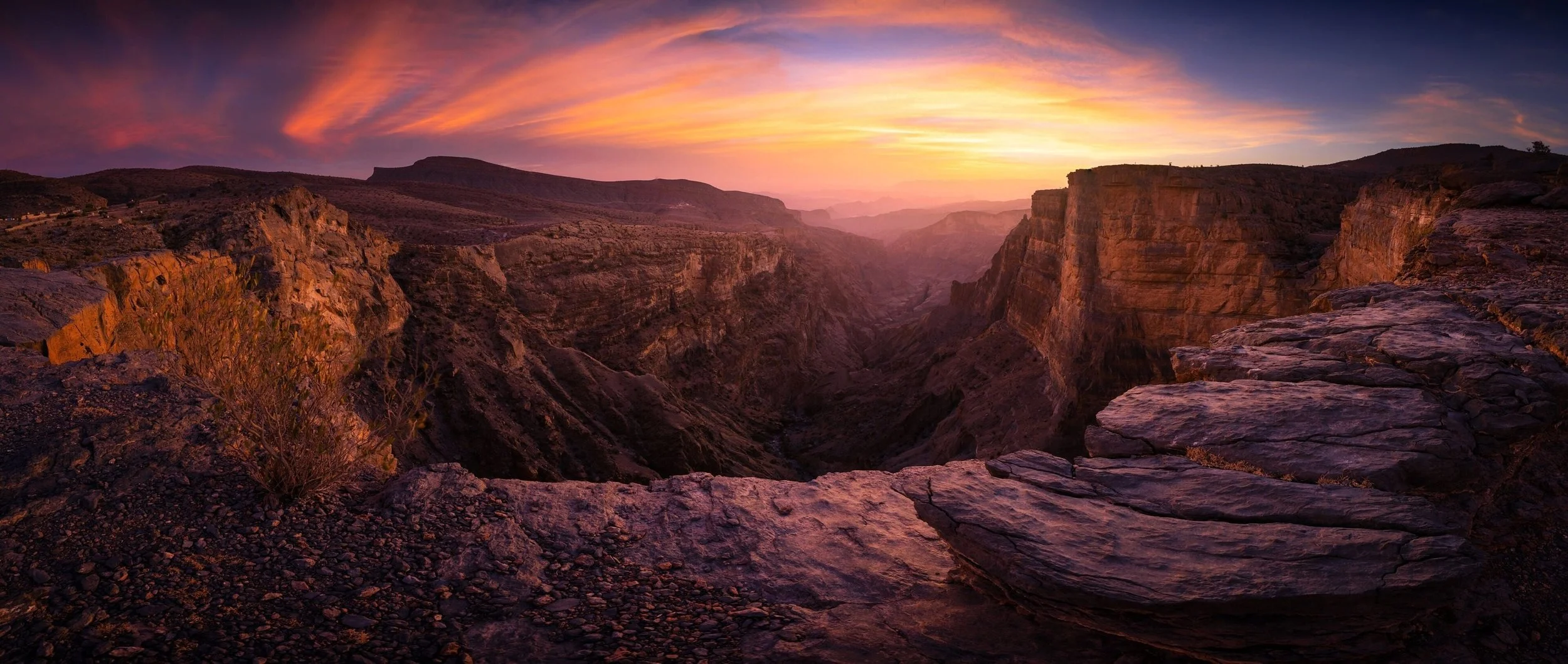 Sunset over a deep canyon with rocky cliffs and layered rock formations in a desert landscape.