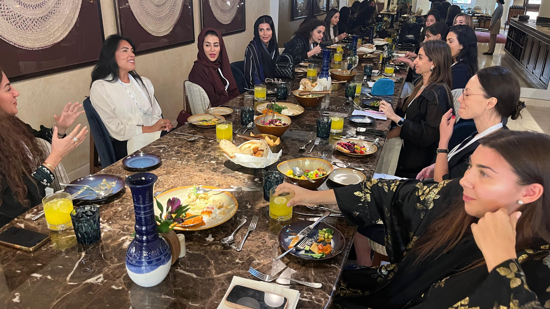 A group of women sitting around a large marble dining table in a restaurant, enjoying a meal and engaging in conversation. The table is decorated with various salads, bread, drinks, and decorative vases.