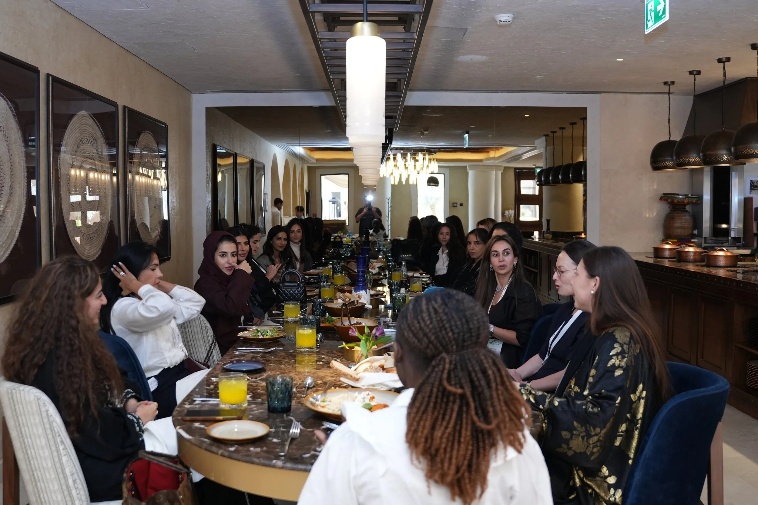 A large group of women seated at a long dining table in a restaurant, engaged in conversation. The table is set with plates of food and glasses of orange juice. The restaurant has modern decor with artwork on the walls and pendant lighting.