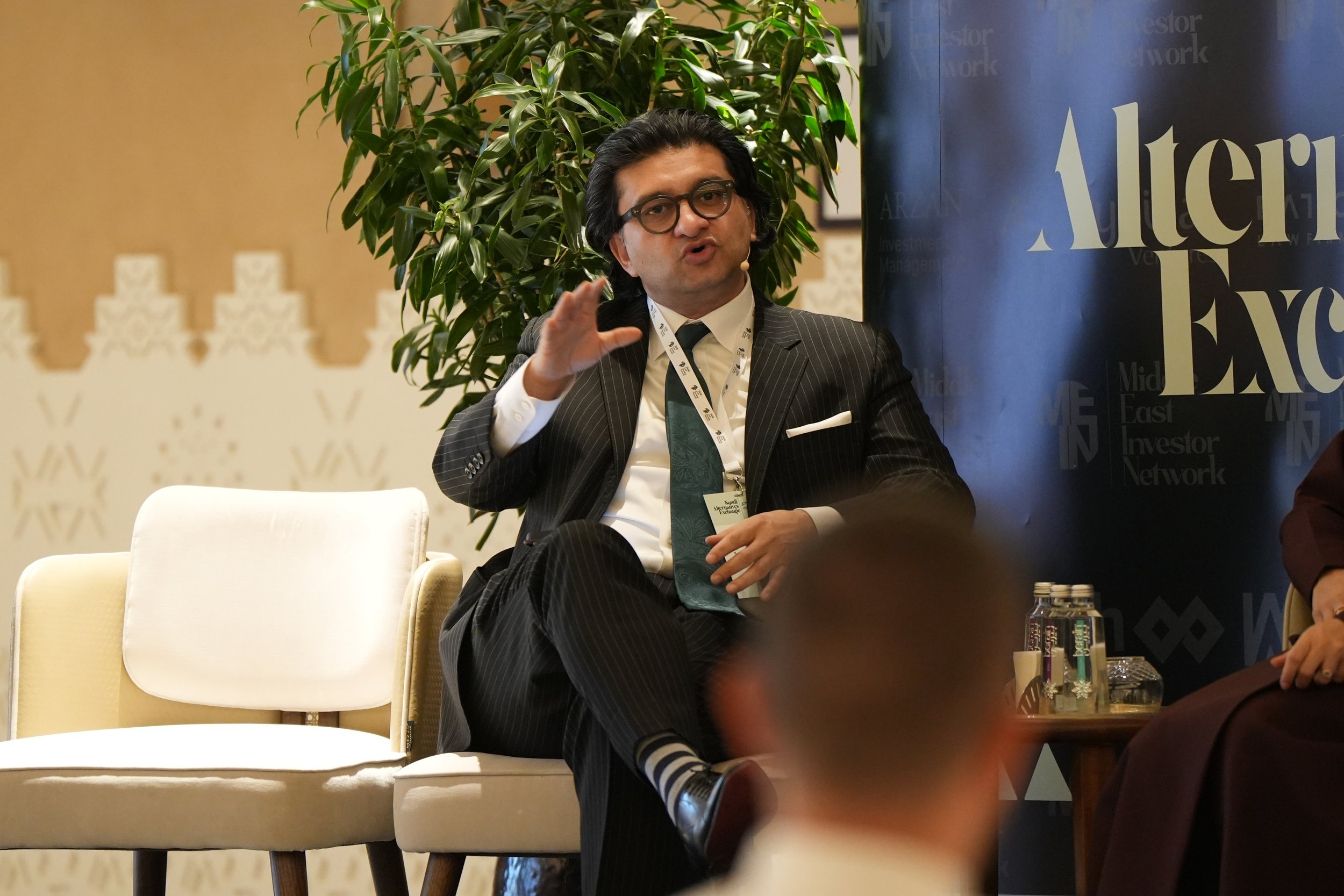 A man with black hair, glasses, and a striped suit is speaking at an event. He is seated with a white lanyard and microphone headset, gesturing with his right hand. Behind him is a large green plant and a banner with the words 'Investor' and 'Network