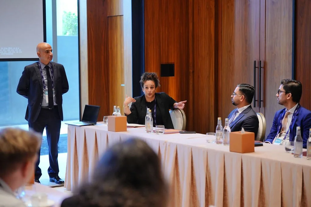 A woman speaking at a conference table with four men listening, in a room with wooden walls and large windows.