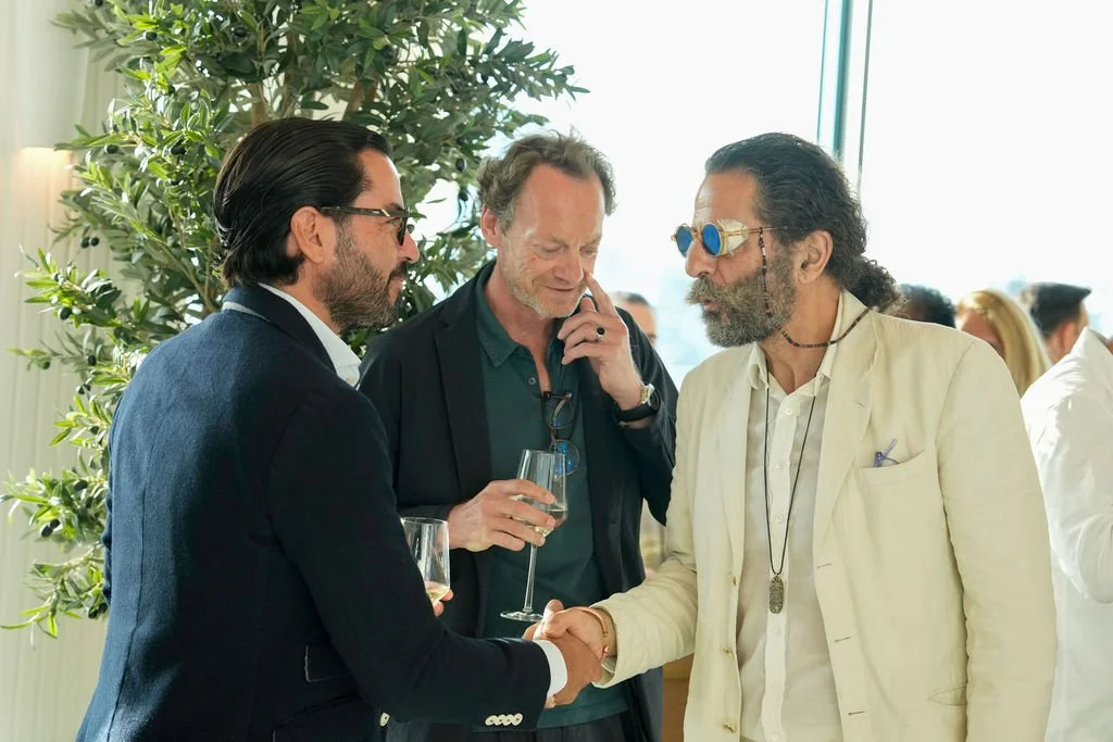 Three men at a social event shaking hands, two holding glasses of wine, in a bright room with a large window and greenplant in the background.