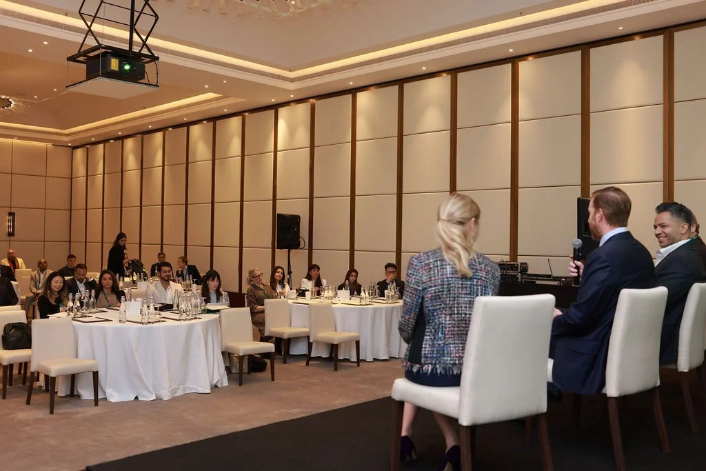 A conference or panel discussion in a large, well-lit banquet hall. Four people sit at the front, with a woman in a plaid blazer speaking into a microphone and three men beside her. The audience, seated at round tables, listens attentively.