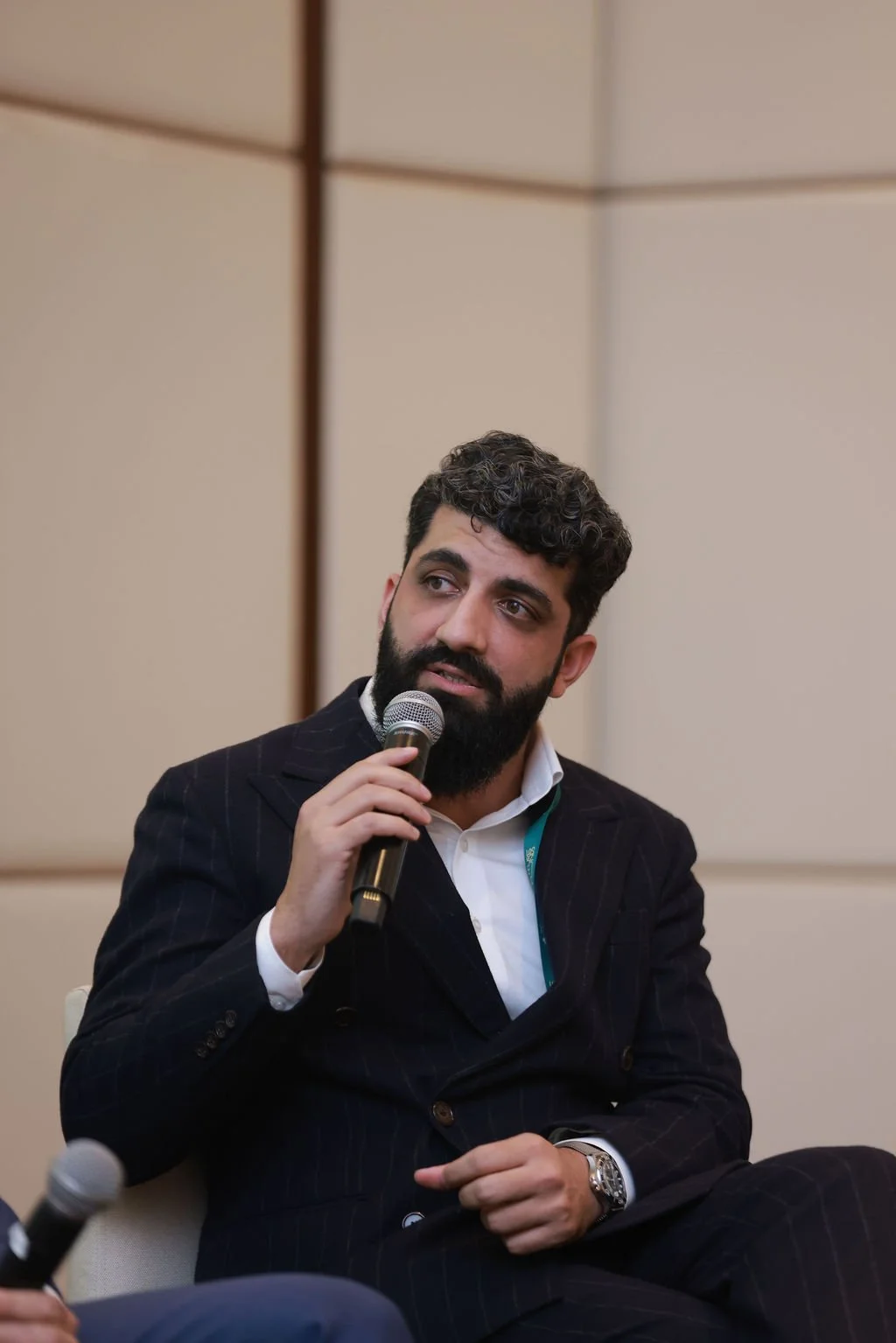 A man with dark, curly hair and a beard, wearing a dark suit and white shirt, holding a microphone and speaking at an indoor event.