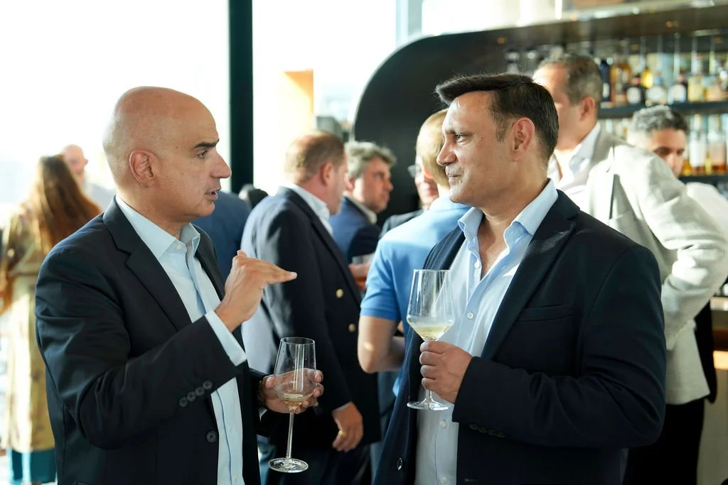 Two men in suits talking and holding wine glasses at a social gathering, with a bar in the background and other people mingling.