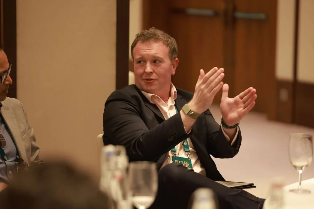 A man in a dark suit and light shirt sitting at a table, clapping with his hands, in a conference room setting.