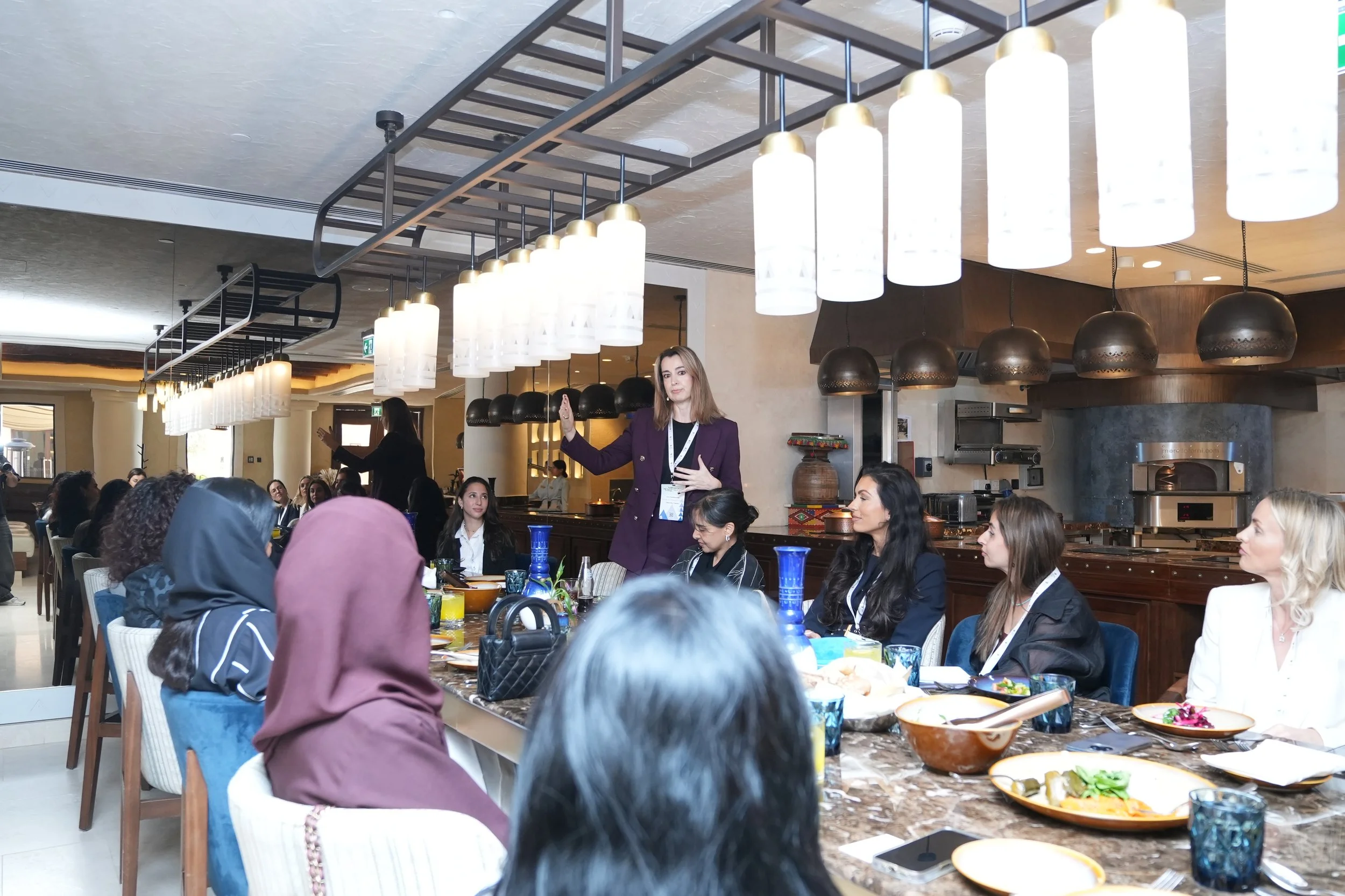 A woman appears to be giving a speech or presentation at a large, diverse banquet or conference with many people seated around tables, and a decorative hanging light fixture overhead.