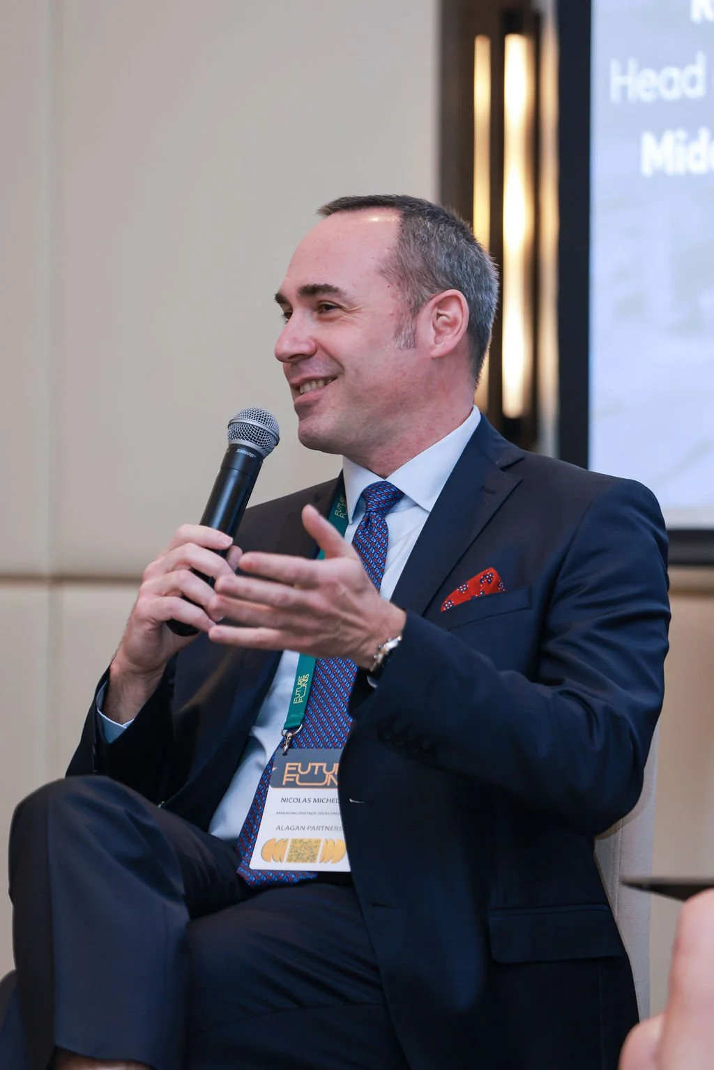 A man in a dark suit, white shirt, and blue patterned tie is sitting on a chair, holding a microphone, and speaking at a conference or seminar. He has a name badge hanging around his neck. There is a large screen with presentation content in the back