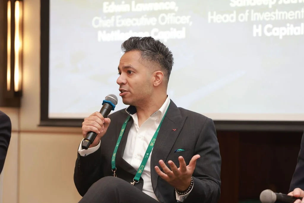 A man in a dark suit with a white shirt, speaking into a microphone during a panel or presentation at a conference or event.