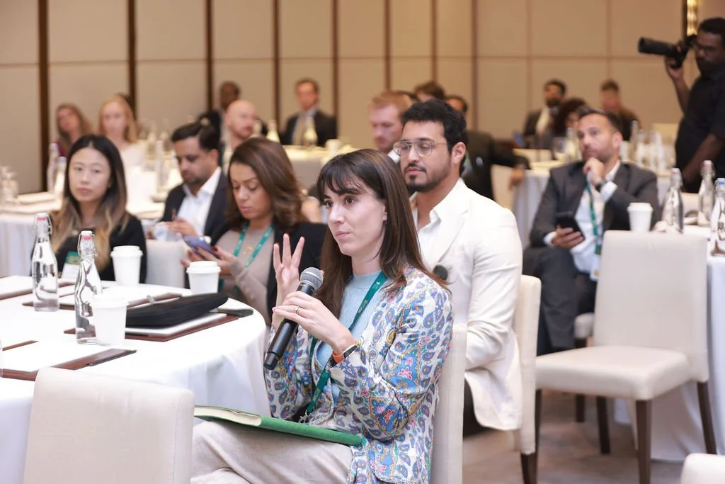 A woman holding a microphone and raising her hand during a conference or seminar, with other attendees seated at tables in the background.