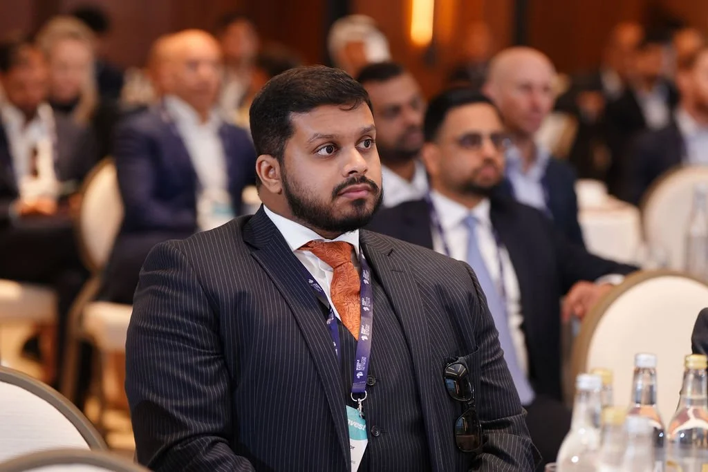 A man in a suit and tie sitting at a conference, listening attentively, with other professionals in the background.