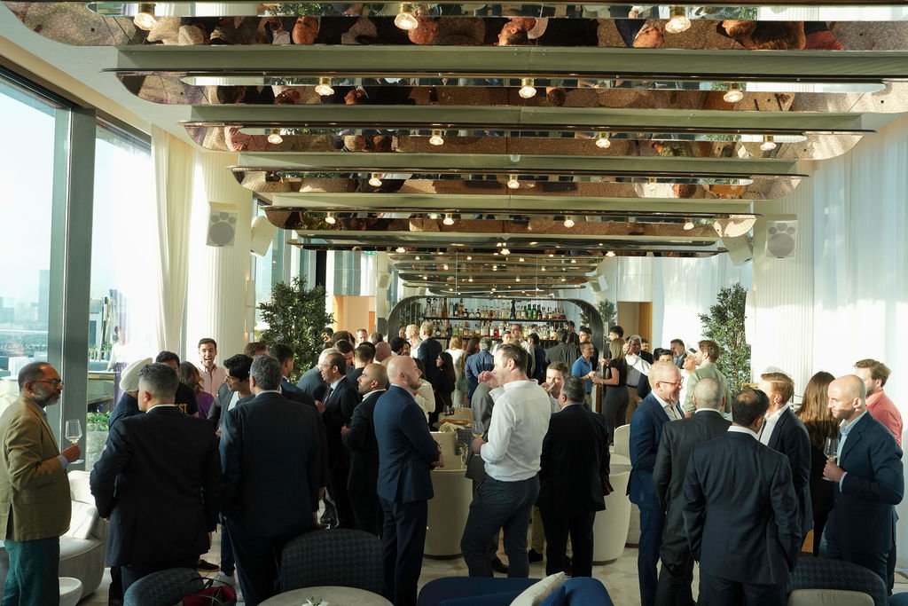 A busy networking event or conference with many people in business attire socializing in a modern, well-lit room with floor-to-ceiling windows and a bar in the background.