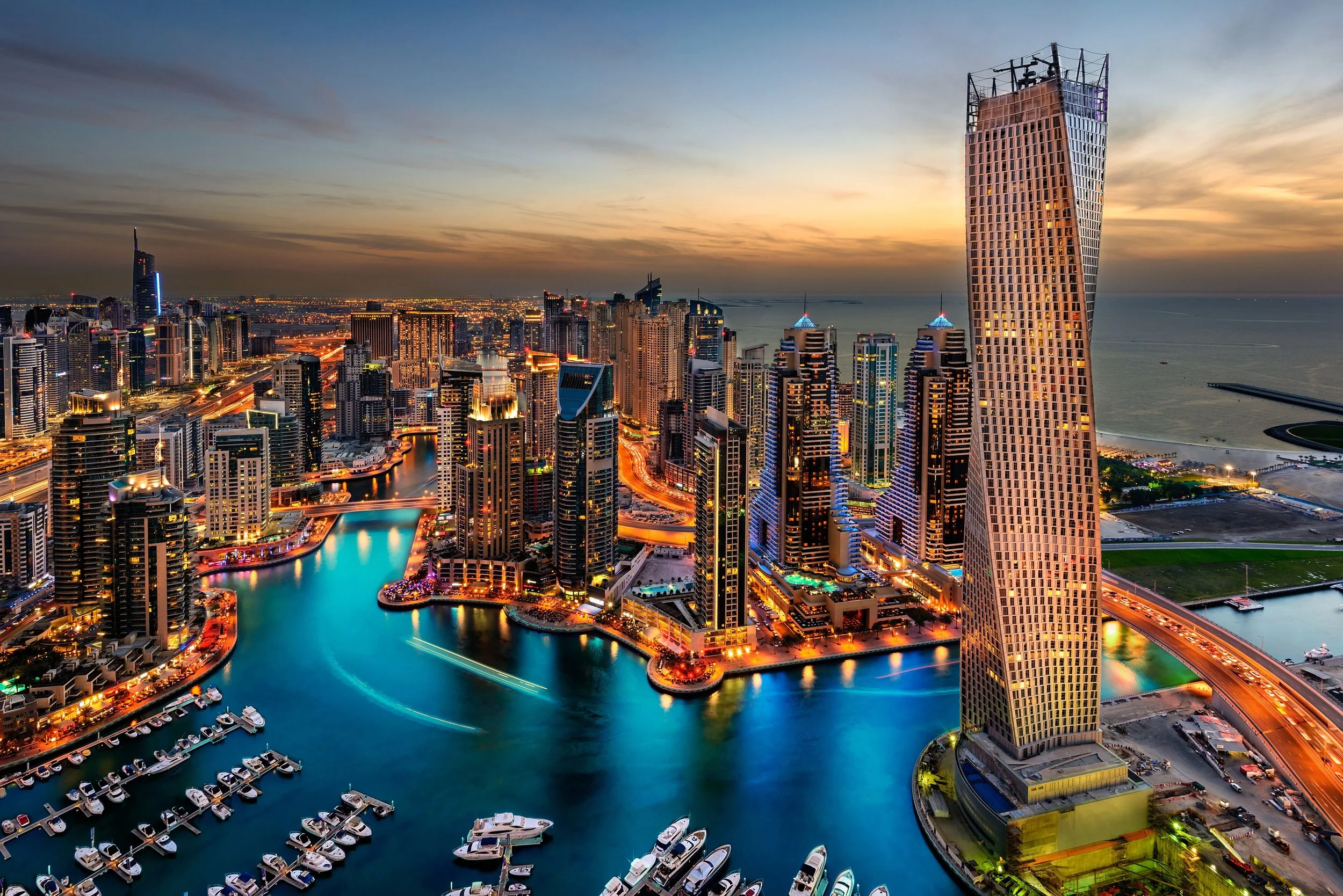 A city skyline at sunset featuring tall modern skyscrapers, with a prominent high-rise building with a distinctive slanted design on the right. The city is illuminated with lights, and a body of water with boats and yachts is visible in the foreground.