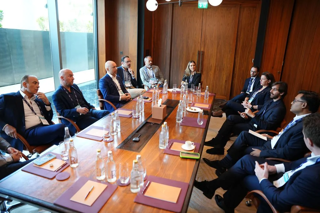 Business meeting with professionals sitting on both sides of a long wooden conference table in a room with wooden walls, large windows, and natural lighting.