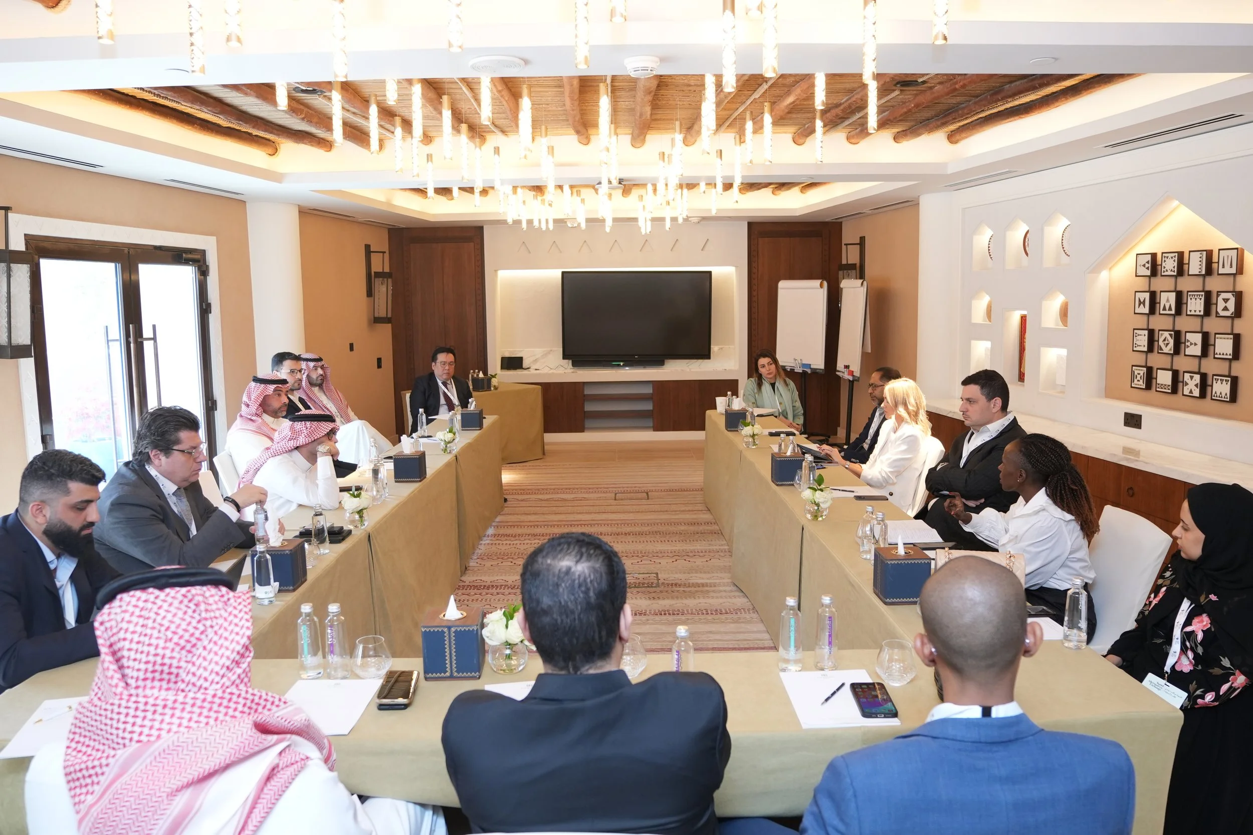 A diverse group of people attending a business meeting or conference in a modern conference room with a wooden ceiling, decorative lighting, and framed artwork on the walls. Attendees are seated around a U-shaped arrangement of tables, some with lapt