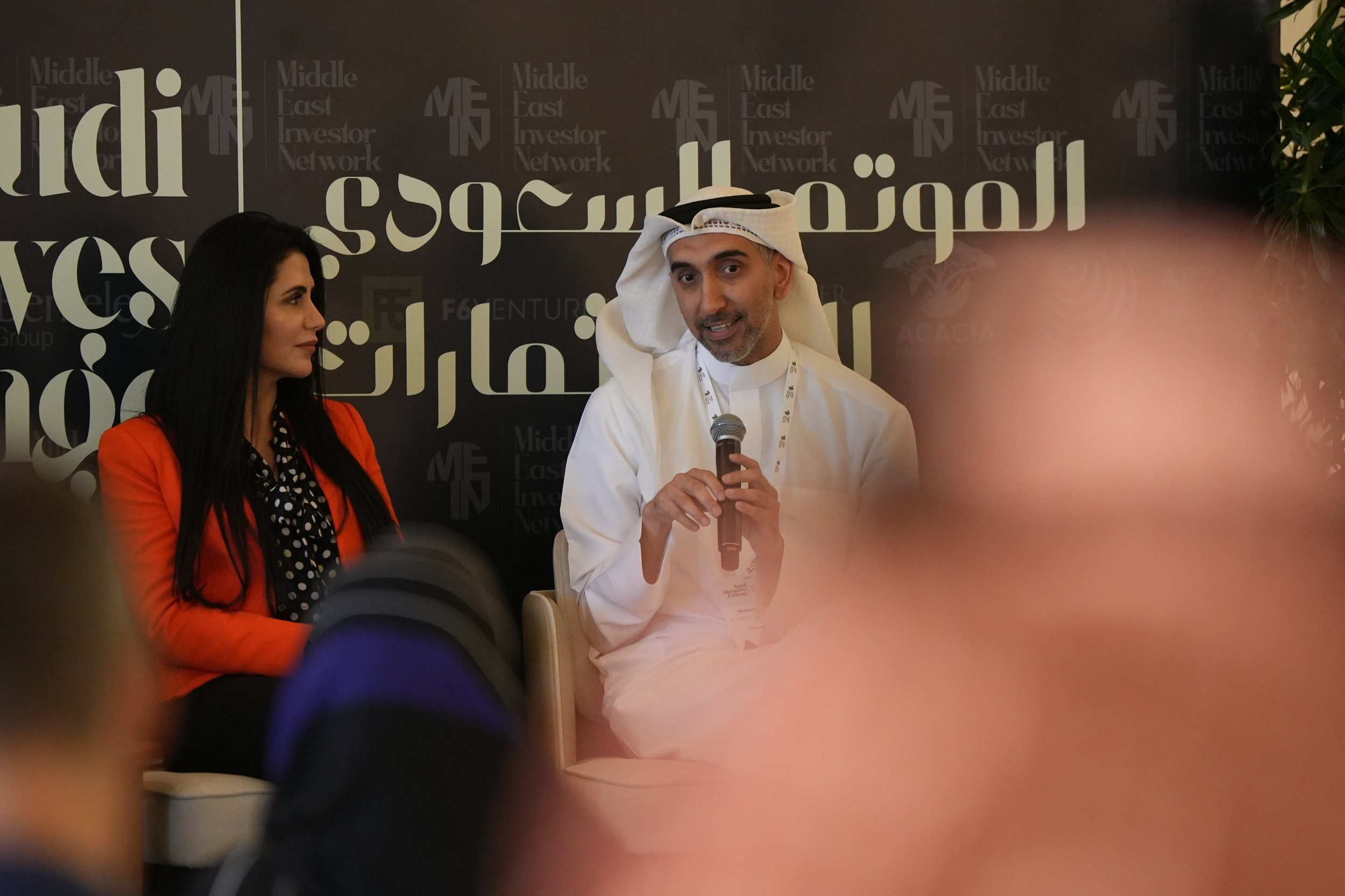 A man in traditional Middle Eastern attire speaking into a microphone at an event, with a woman in a bright orange blazer sitting next to him, in front of a black backdrop with Arabic and English text that reads "Middle East Investor Network."