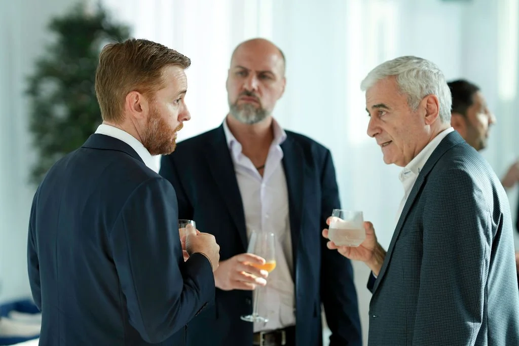 Three men in business attire having a conversation at a social event, holding drinks. Two of them are listening attentively while one appears to be speaking, with a blurred background showing other people and a plant.