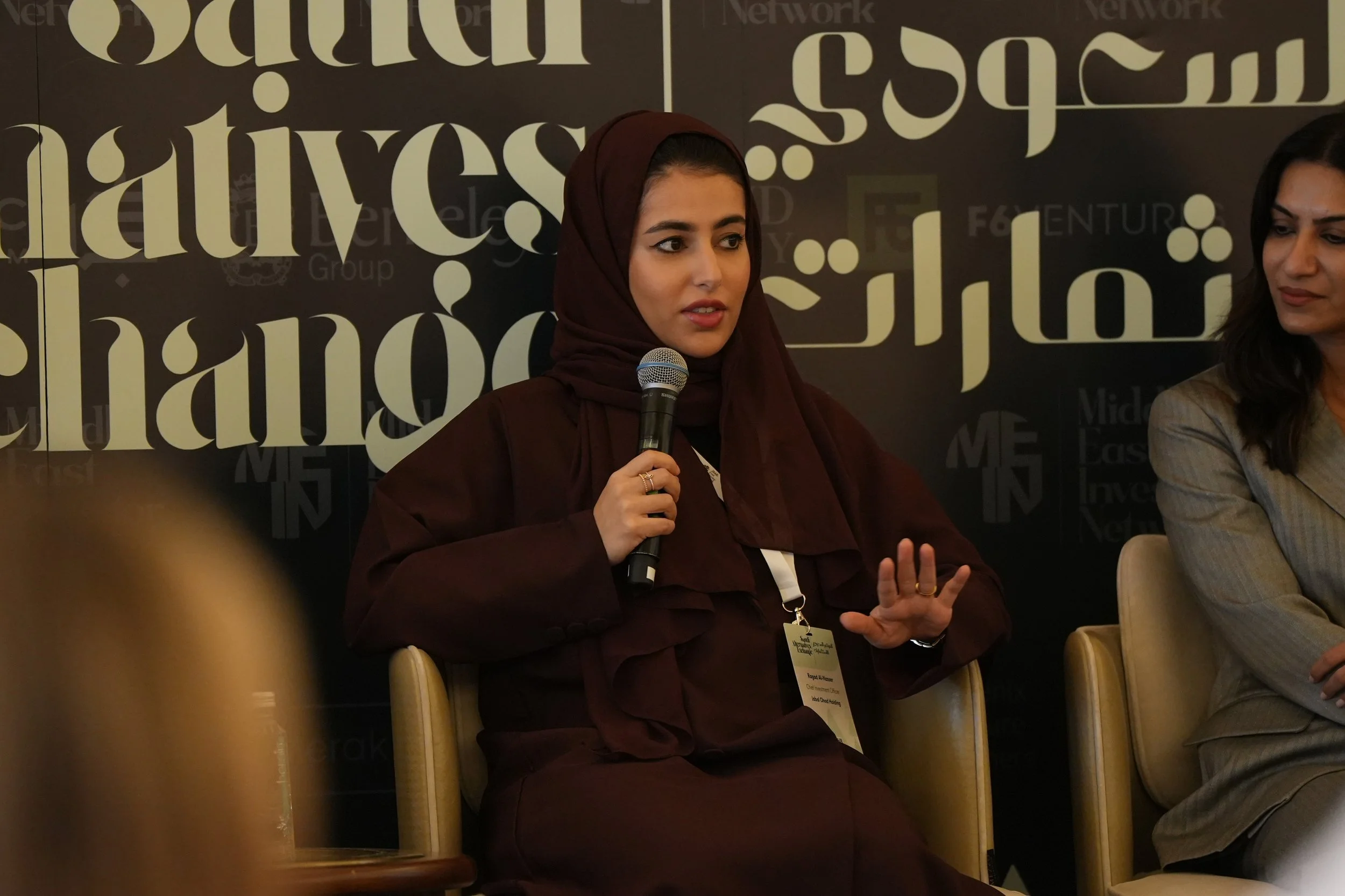 A woman wearing a maroon headscarf and dark clothing speaking into a microphone at an event. She has a name badge hanging around her neck. Another woman is sitting beside her. The background features large stylized text and logos.