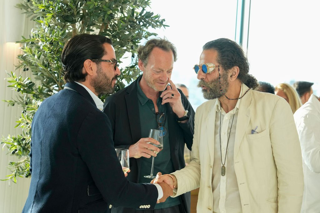 Three men exchanging a handshake at a social gathering, with a woman and other people blurred in the background, and a large potted plant nearby, indoors with bright natural light.