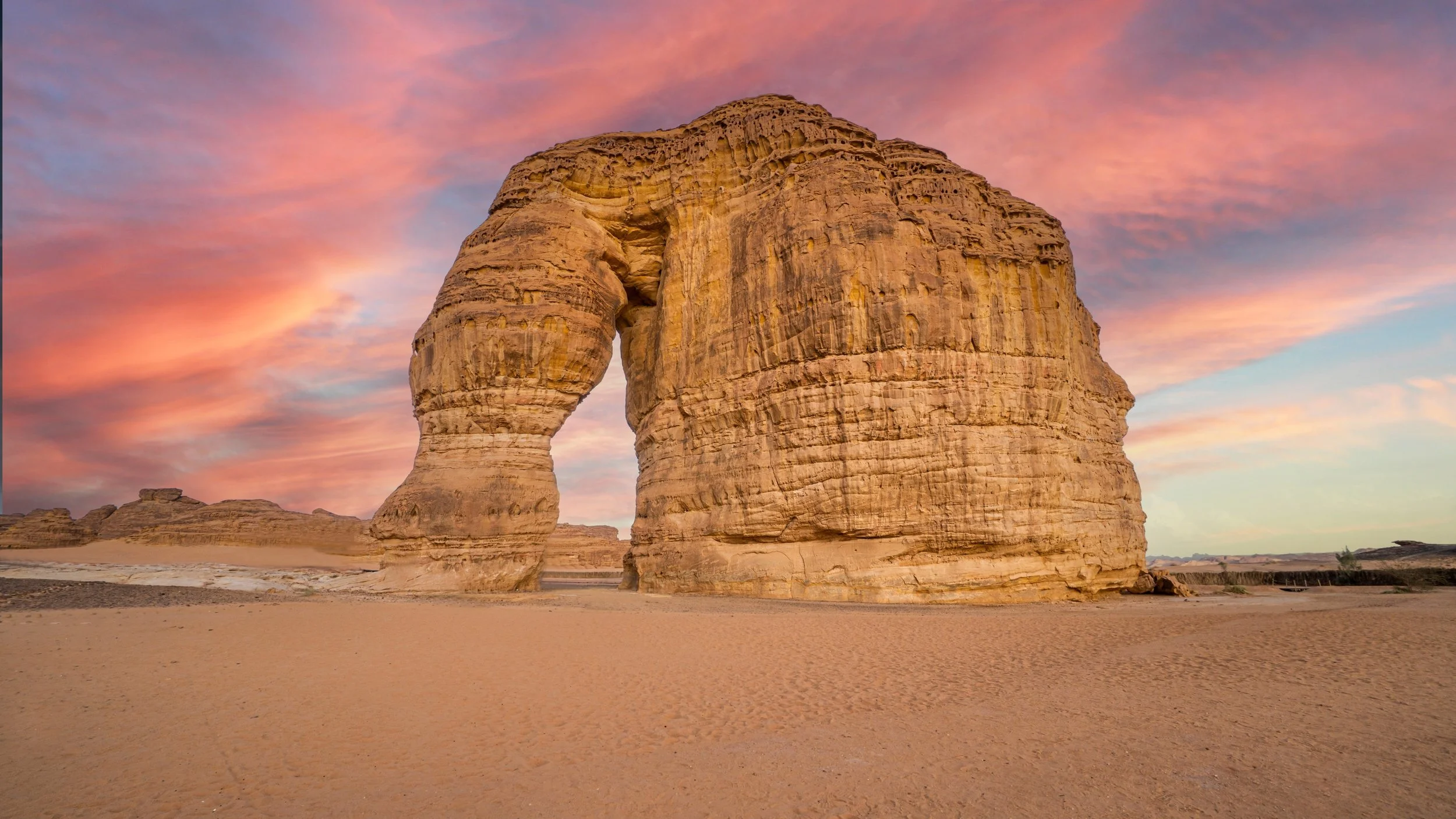 A large, unique rock formation in a desert landscape during sunset with a pink and orange sky.