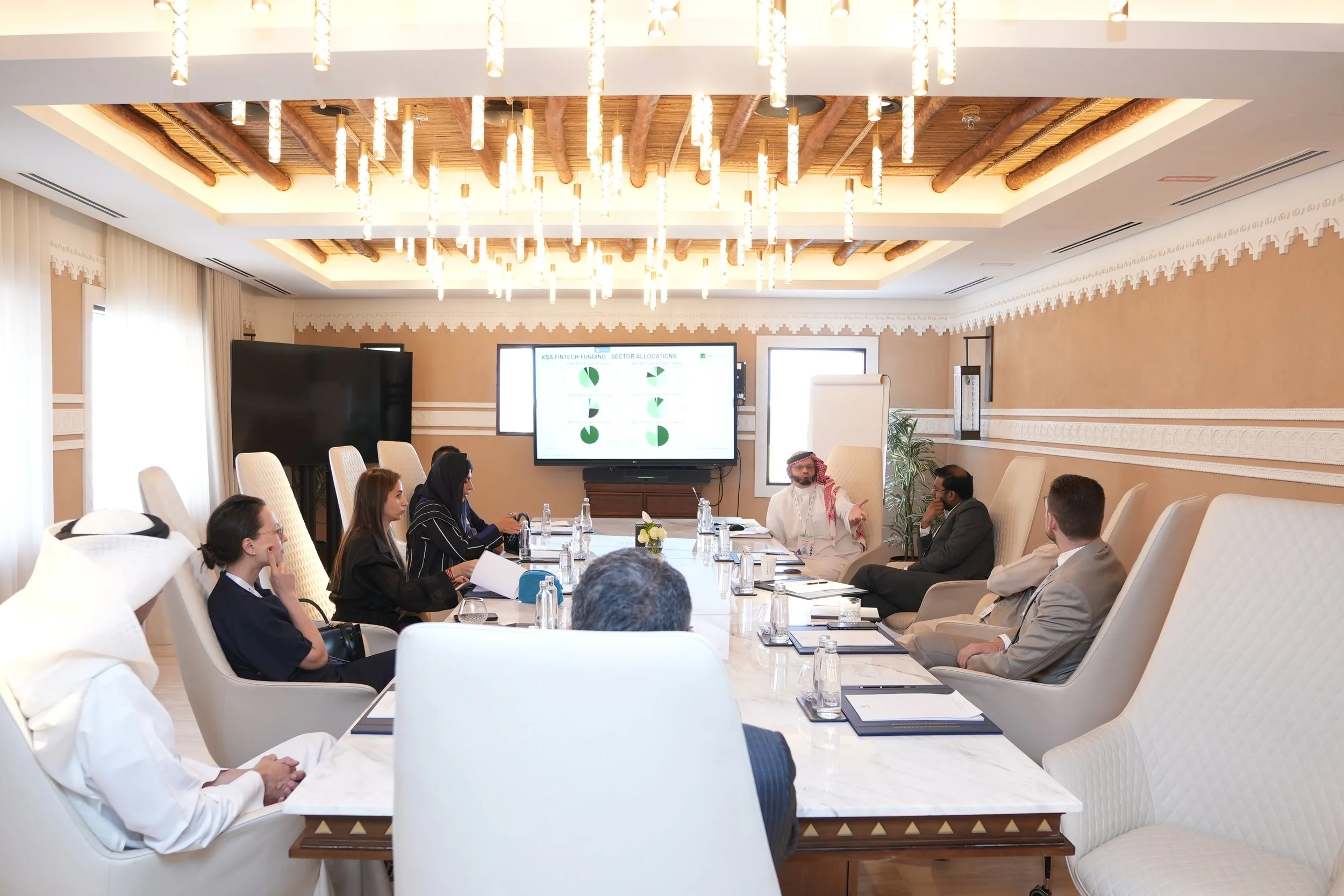 A group of diverse people seated around a large conference table in a well-lit, elegant meeting room. The room has a high ceiling with decorative wooden beams and modern hanging light fixtures. A large screen at the front displays a presentation, and there are a few bottles of water and notebooks on the table.