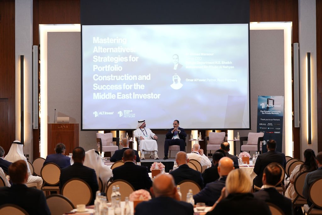 Professional conference with speakers on stage discussing investment strategies, with an audience seated at round tables in a large, well-lit room.