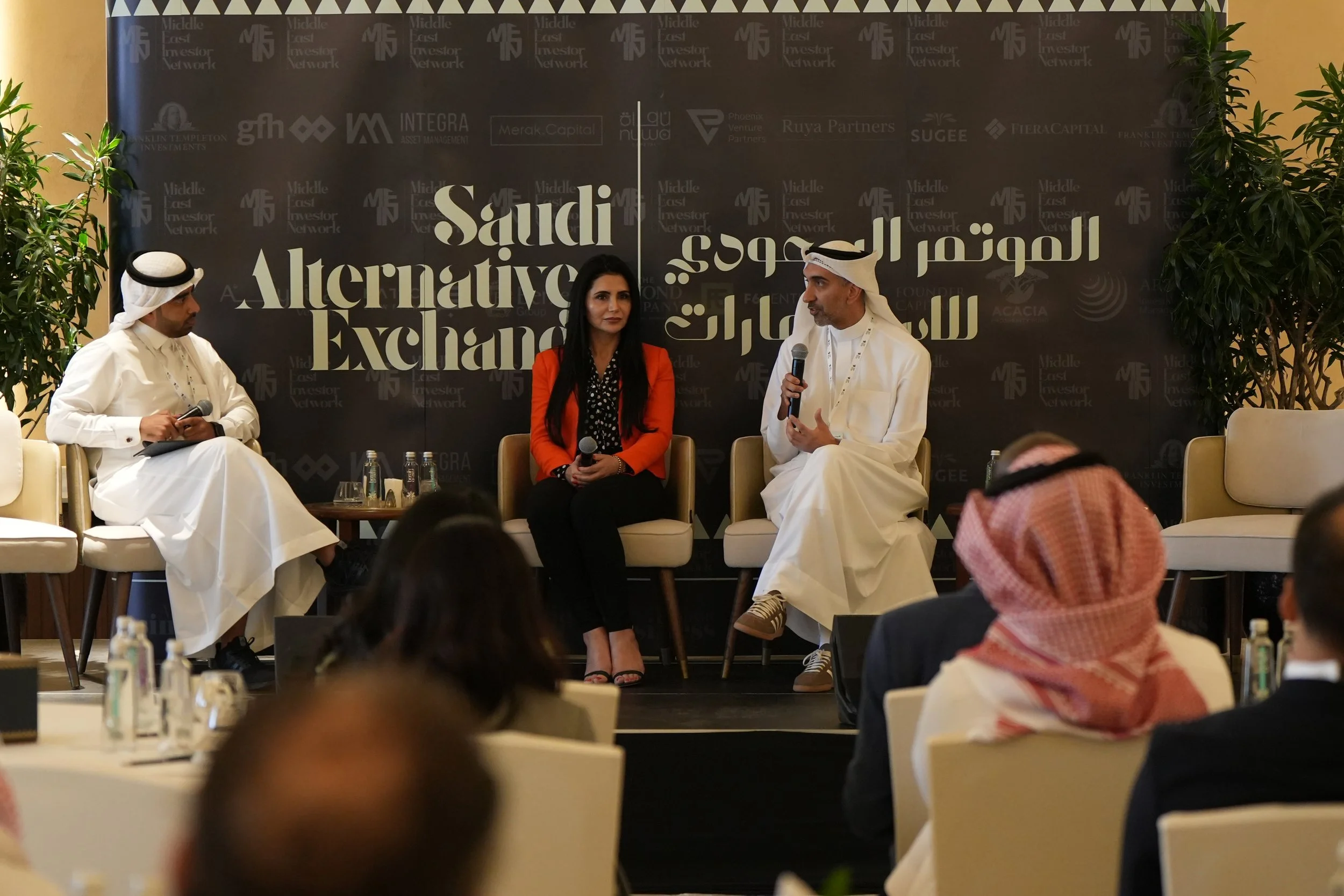 Panel discussion at the Saudi Alternative Exchange event, with three individuals seated on stage, two men wearing traditional Gulf attire and a woman in a black and red outfit, against a dark backdrop with logos and Arabic text.