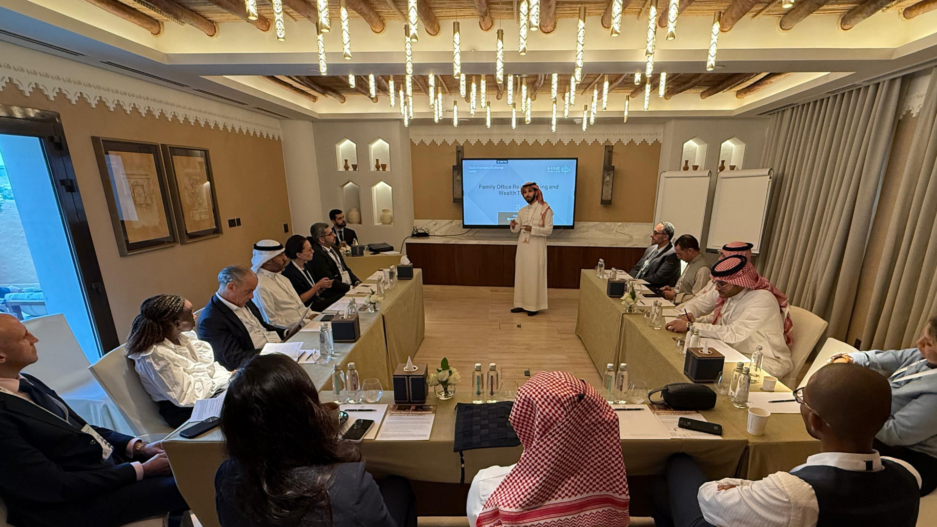 A professional conference room with participants seated around rectangular tables, facing a presenter at the front. The presenter is a man in traditional Middle Eastern attire, speaking to the group. A large screen behind him displays a presentation 