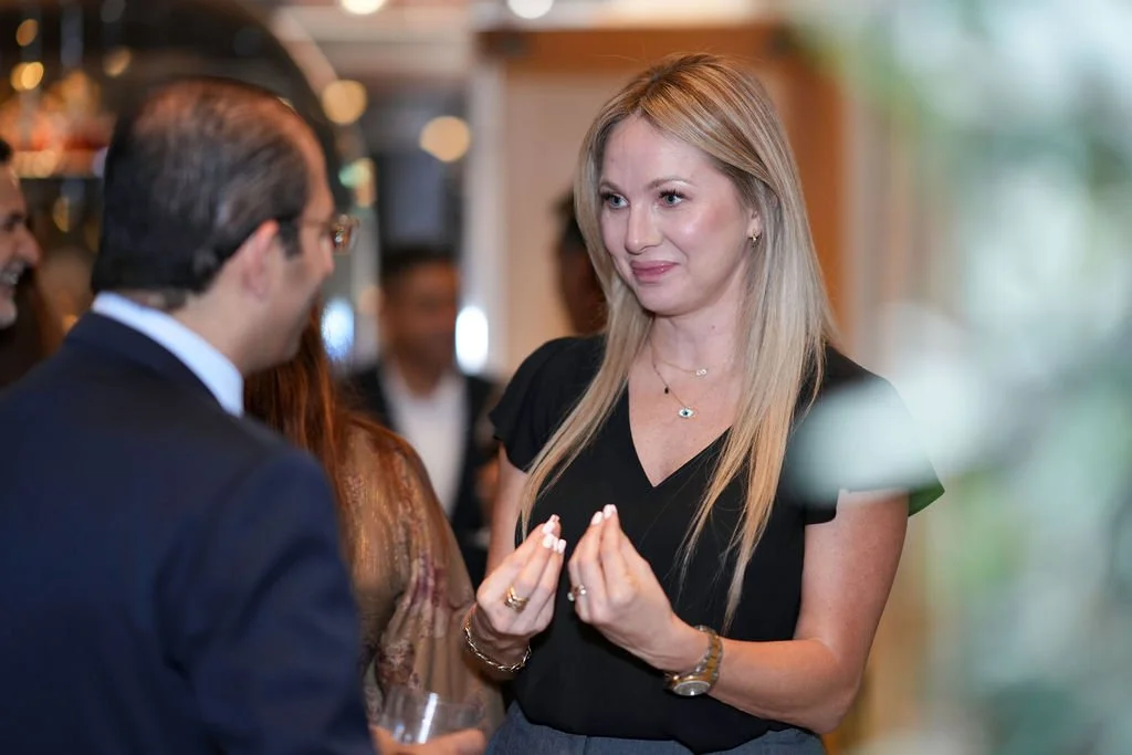 A woman with long blonde hair wearing a black top, jewelry, and a watch, chatting with a man in a suit at a social event.