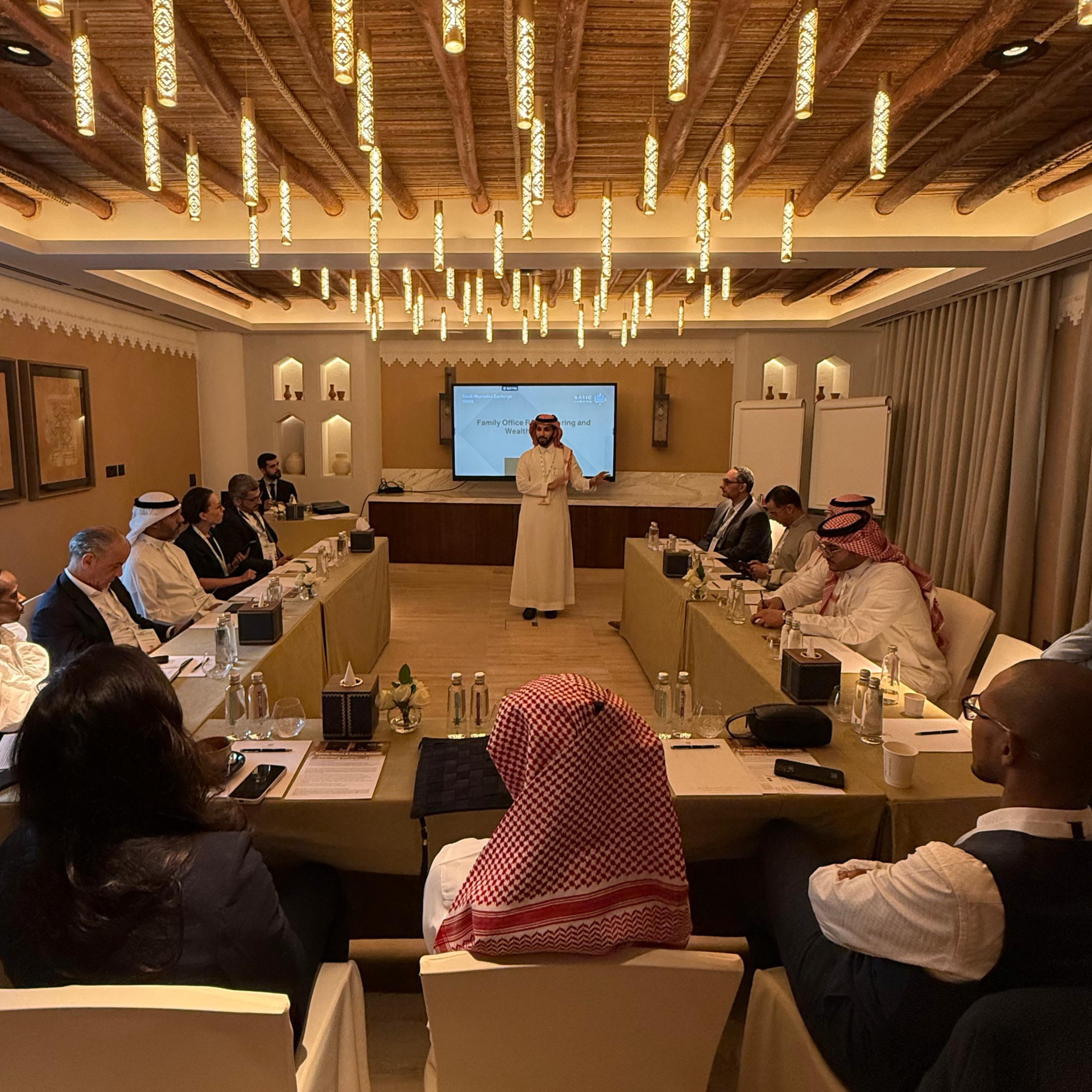 A conference or meeting room with people seated around a large U-shaped table, listening to a speaker standing at the front. The room features ornate, wood-beamed ceiling with hanging lights, decorative wall niches with vases, and a large television screen displaying a presentation. Some attendees are dressed in traditional Middle Eastern attire, such as white kanduras and headscarves, while others wear business suits. The atmosphere appears professional and formal.