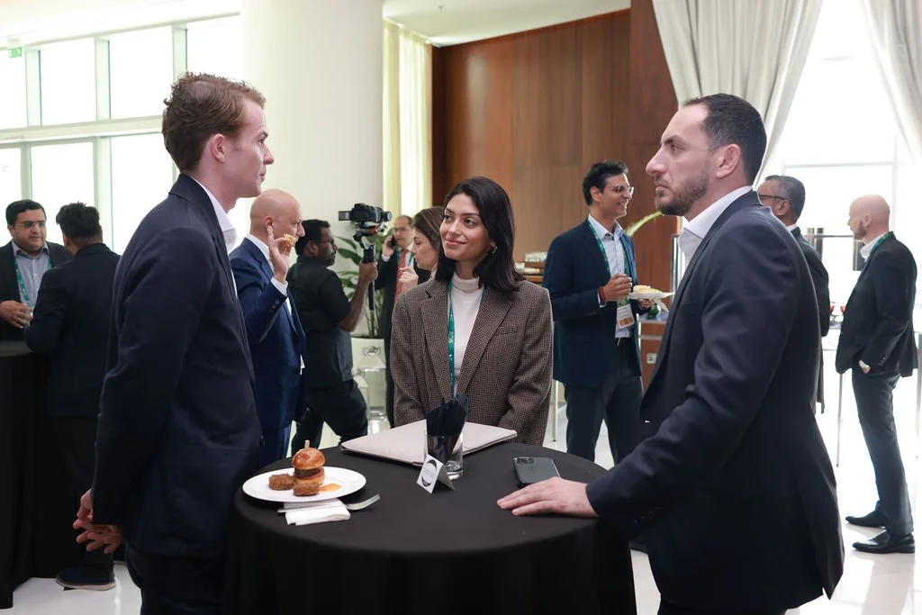 Business professionals engaging in conversation at a networking event, standing around a high table with snacks and a notepad, in a well-lit conference room.