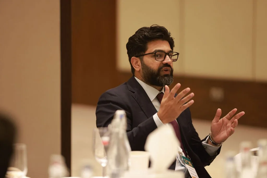 A man with dark hair, beard, and glasses wearing a black suit and white shirt, sitting at a table during a conference or meeting, gesturing with his hands.
