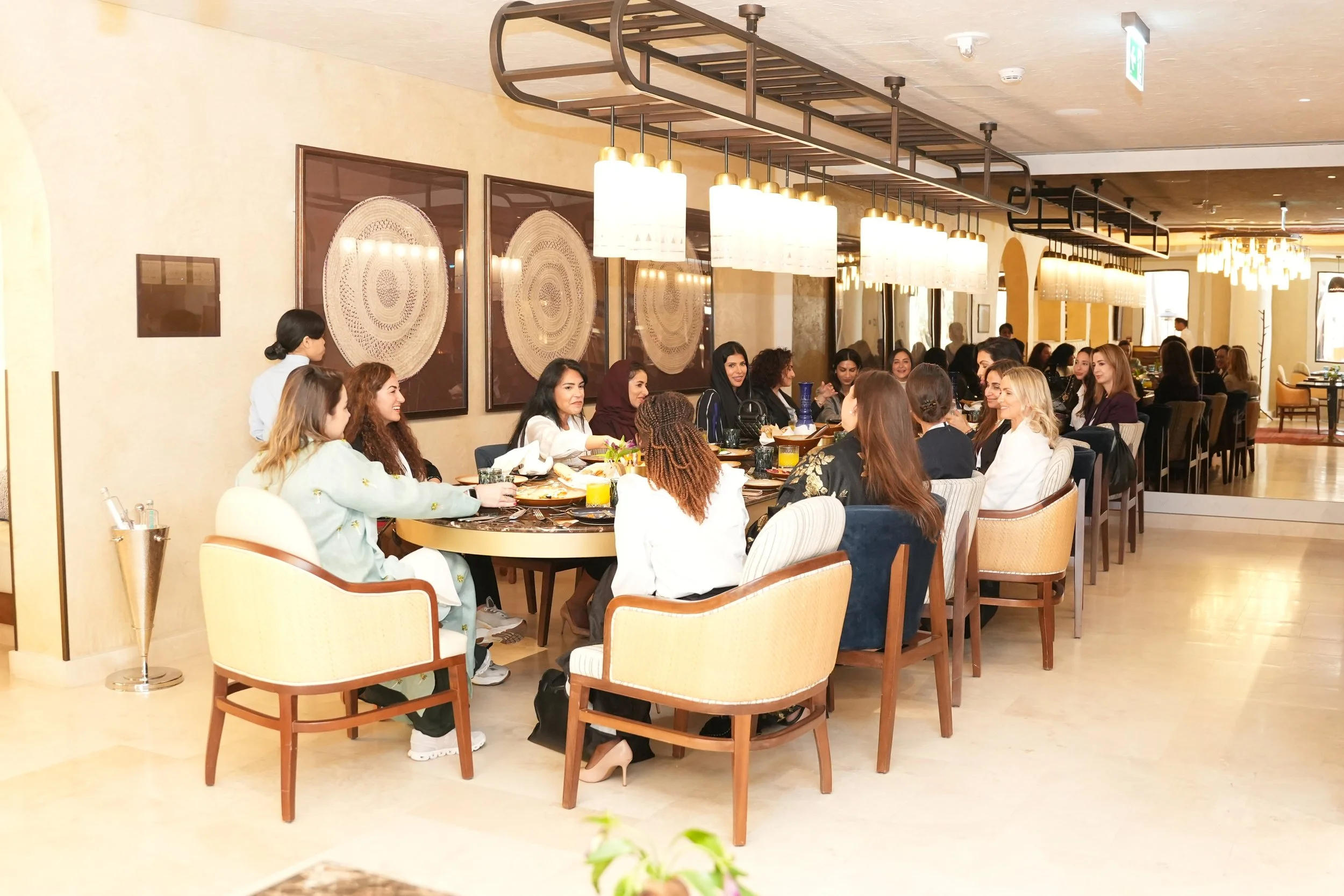 Inside a restaurant with a large group of women seated at tables, engaging in conversation. The decor features modern lighting fixtures and artwork on the wall.