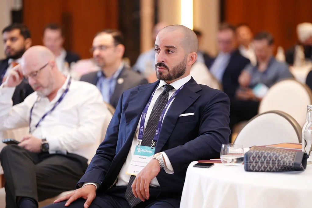 A man with a shaved head and beard wearing a navy suit, white shirt, and patterned tie sitting at a conference table, attentively listening. Other conference attendees are visible behind him, some taking notes or using devices.