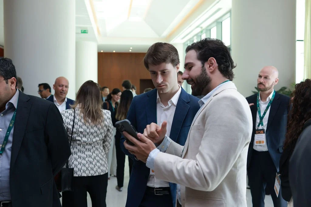 Two men in suits are looking at a smartphone together at a business conference, with other attendees in the background.
