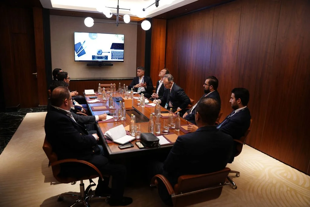 Business meeting with nine people seated around a conference table in a wood-paneled room. There are bottles of water, notebooks, and electronic devices on the table. A large screen is mounted on the wall at the front showing presentation slides with