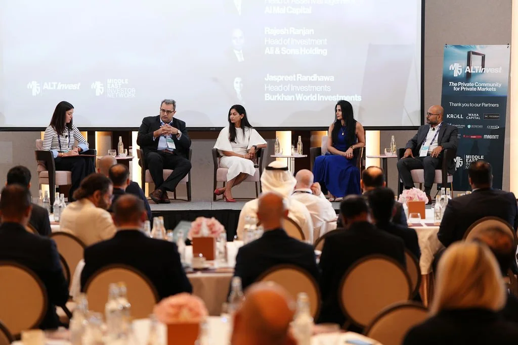 Panel discussion at a conference with five speakers on stage, seated in armchairs, in front of an audience in a ballroom setting. A large screen behind them displays text and logo.