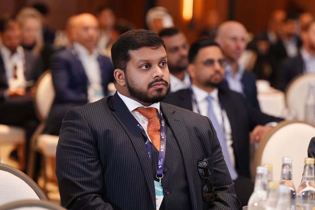 Man in a dark pinstripe suit, white shirt, and orange tie attending a conference with other business professionals.