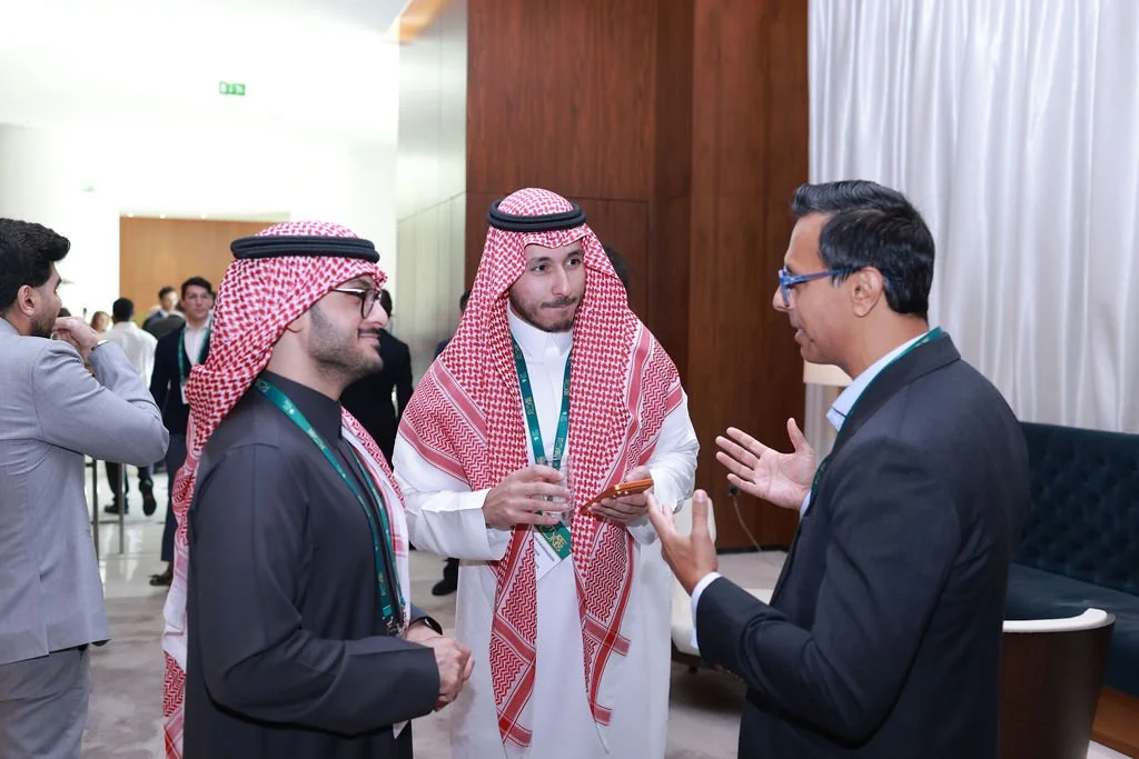 Three men are engaged in a conversation in a professional setting, two wearing traditional Middle Eastern attire with head coverings, and one in a business suit and glasses. Other people are visible in the background.