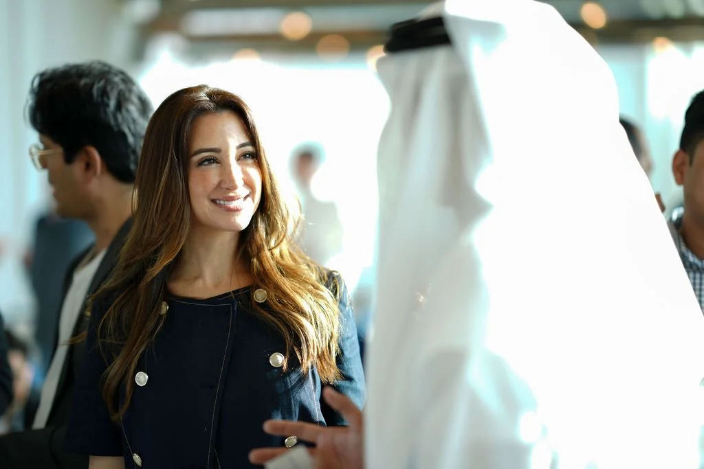 A woman with long brown hair smiling and talking to a man dressed in traditional Middle Eastern attire at a formal event.