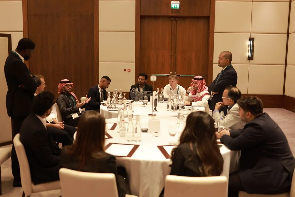 A diverse group of people seated around a large round table in a conference room, engaging in a discussion, with a man standing and speaking to the group.