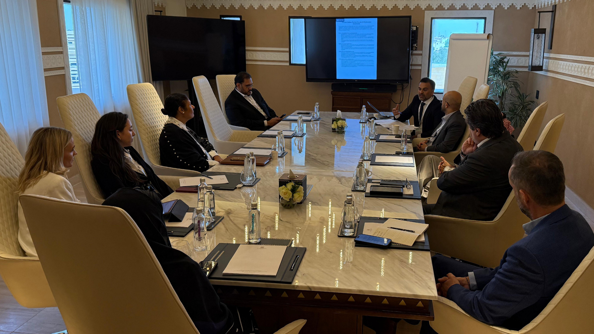 A formal business meeting with ten people seated around a large marble conference table in a well-lit room, with a large screen displaying a document at the front.