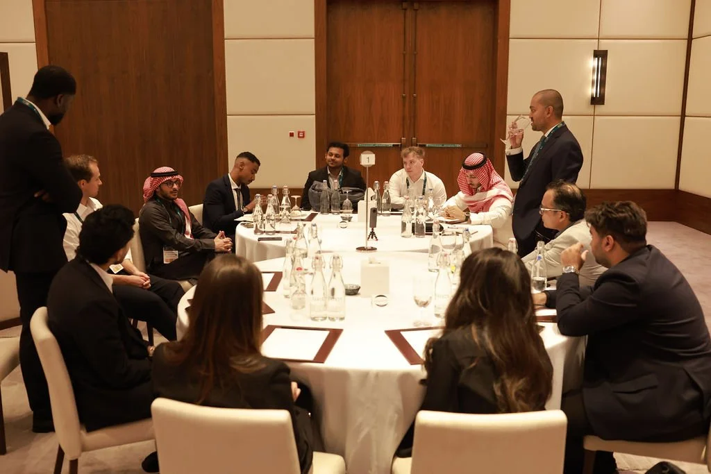 A group of diverse professionals sitting around a large, round conference table in a formal setting, with one man standing and speaking, and another man drinking water.