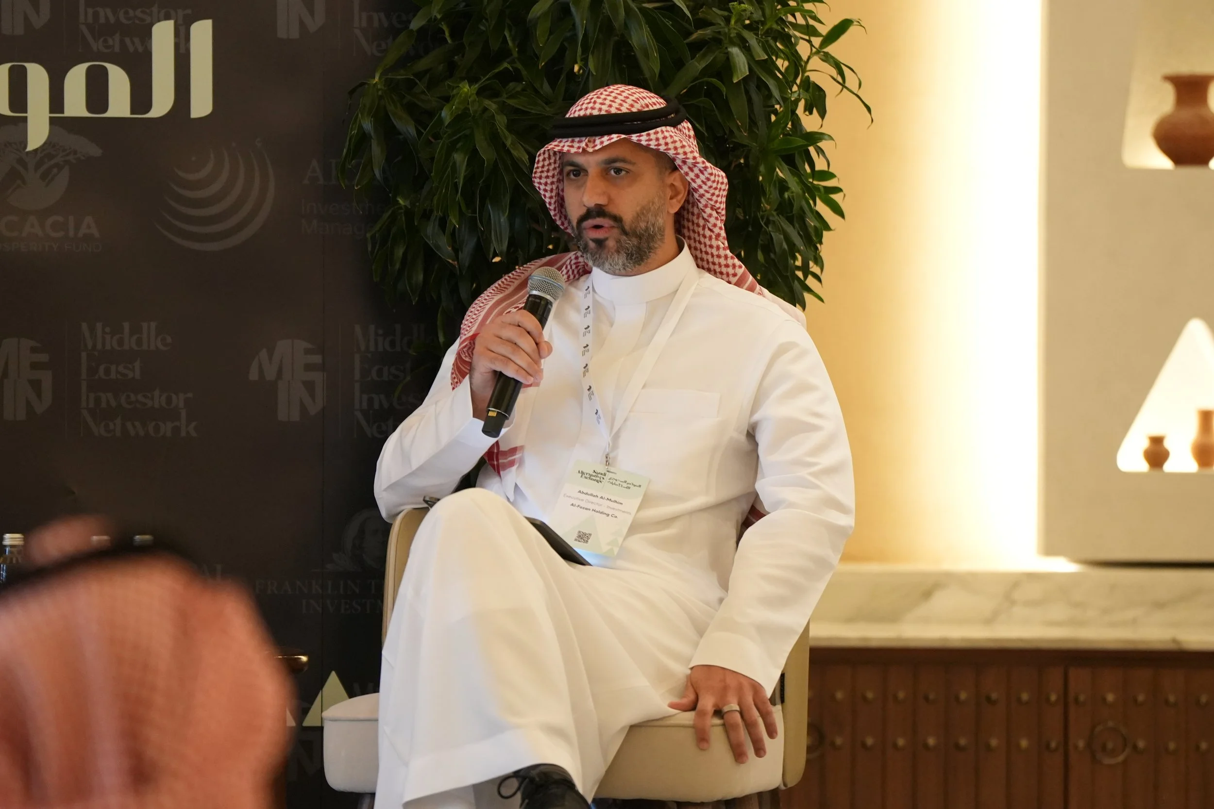 A man wearing traditional Middle Eastern attire, including a red checkered shemagh and white thobe, is speaking into a microphone while seated on a beige chair at a conference or event with a backdrop displaying the logos of Middle East Investor Netw