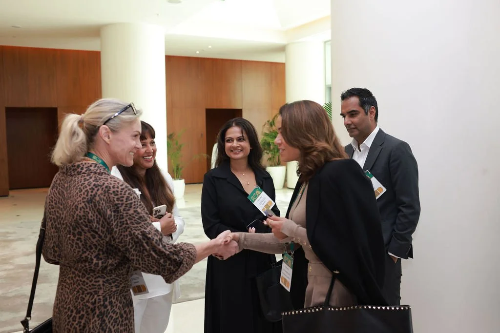 A professional networking event where five diverse individuals are engaged in a conversation, with two women shaking hands while the others observe and smile.