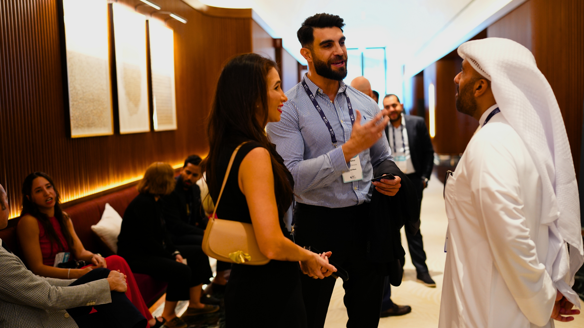 Three people are standing and talking at a conference or professional event, with several seated attendees in the background.