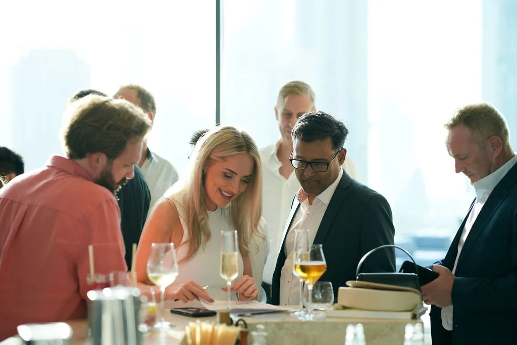 Business professionals gathered around a bar counter, looking at a smartphone with drinks on the counter.