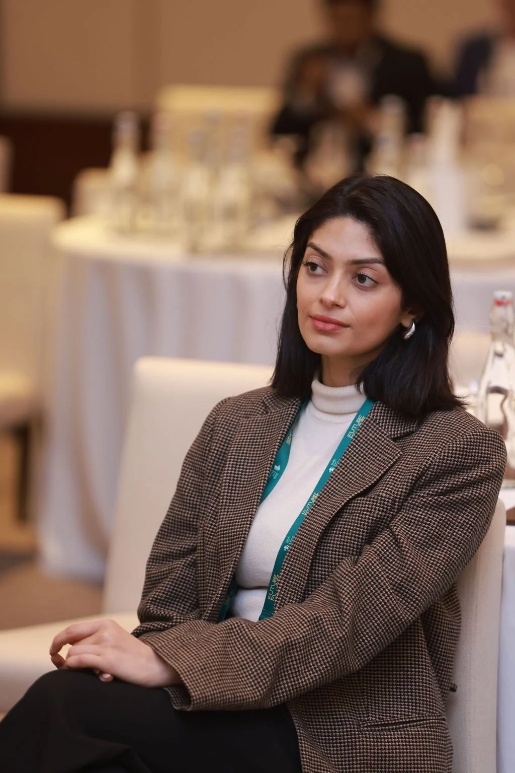 A woman with black hair and hoop earrings sitting in a conference room or banquet hall with round tables and glasses in the background.