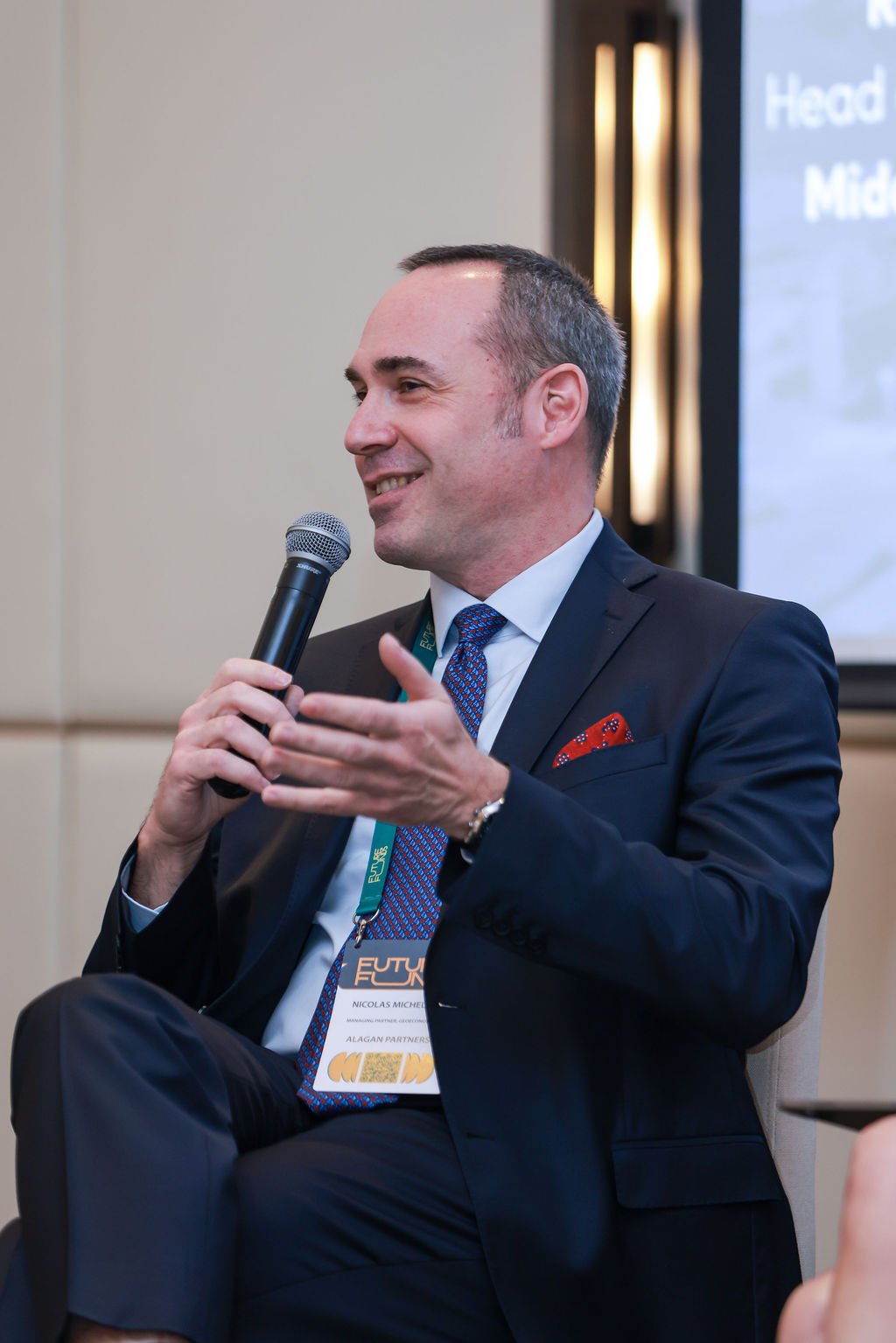 A man in a navy suit, white shirt, and red patterned tie speaking into a microphone during a conference. He's wearing a conference badge around his neck.