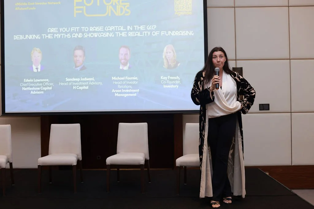A woman standing on a stage holding a microphone, giving a presentation at a conference. Behind her is a large screen displaying a slide with the title 'Future Funds' and images and names of four speakers.